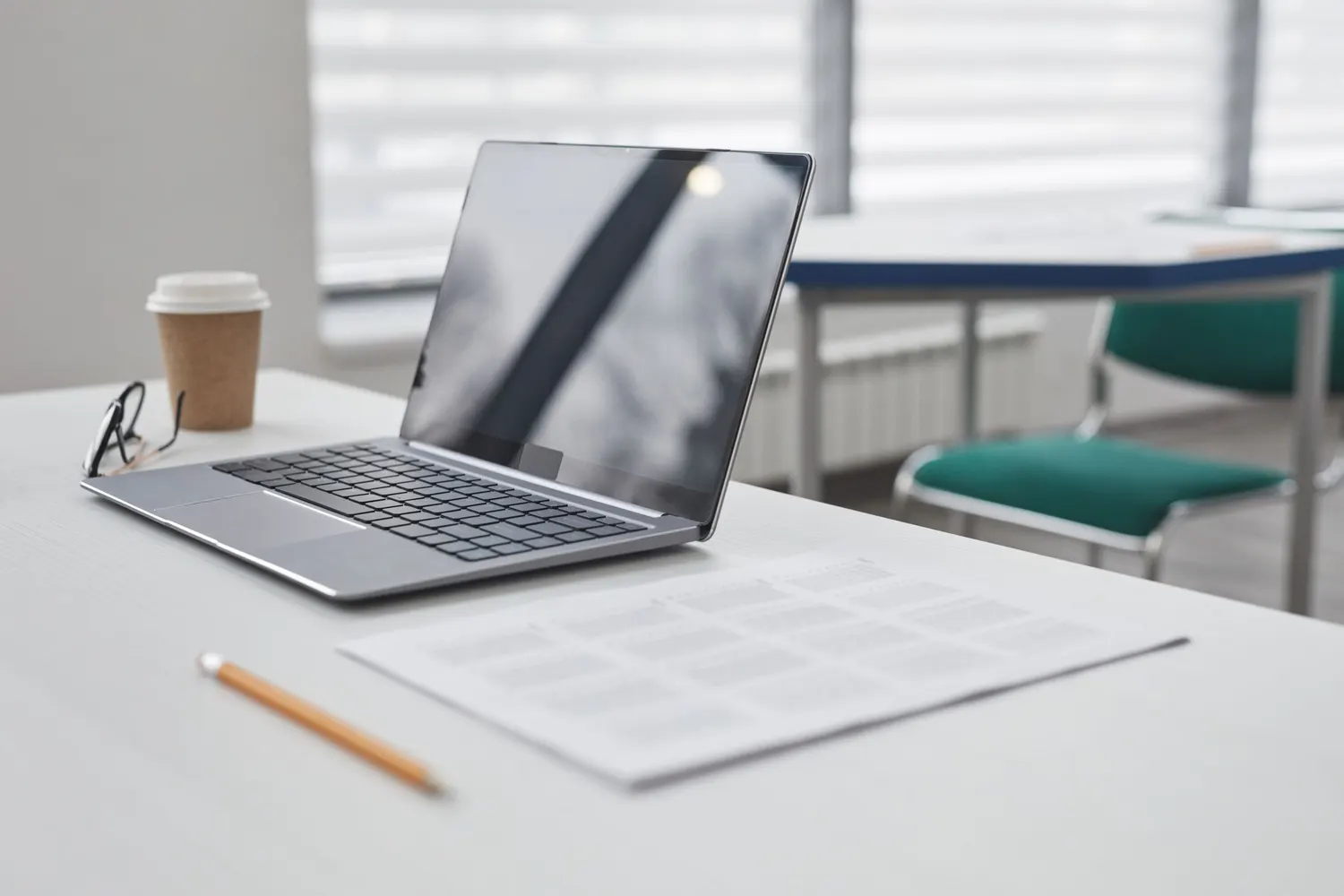 A laptop, a printed form, a pencil and a coffee on a desk by a window