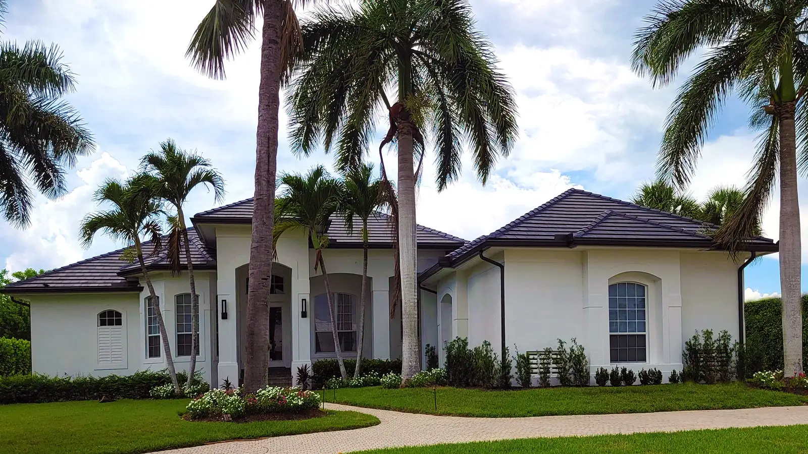 A white single-story stucco house with a tile roof, tall palm trees and a curving paved path