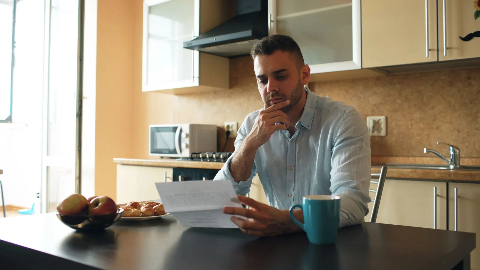 A man at a kitchen table reading a letter, a mug and a bowl of fruit beside him